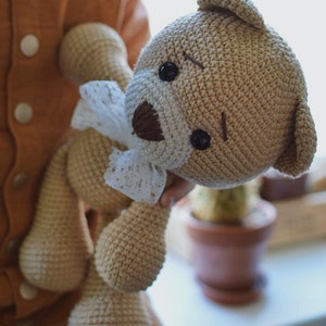 May include: A crocheted teddy bear with brown eyes and a white lace bow. The bear is light brown and has a stitched brown nose.