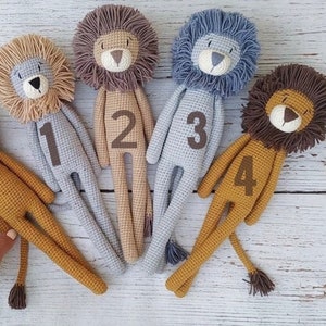 May include: A collection of five handmade crochet lion dolls in various colors. Each lion has a textured mane, a stitched face, and a number from one to four on its body. The lions are lying on a light-colored wooden surface.