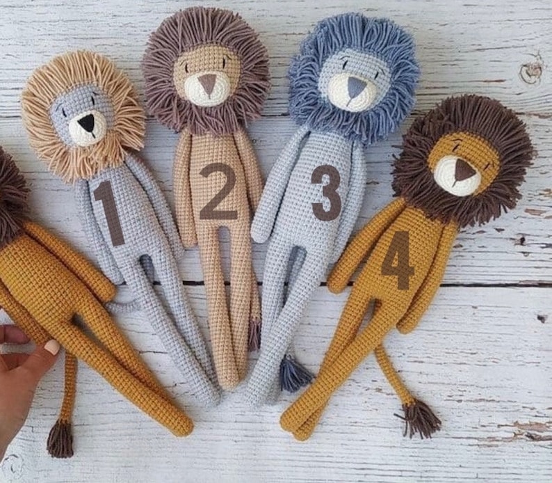 May include: A collection of five handmade crochet lion dolls in various colors. Each lion has a textured mane, a stitched face, and a number from one to four on its body. The lions are lying on a light-colored wooden surface.