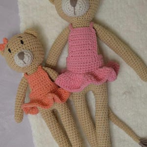 May include: Two handmade crochet teddy bears. One bear wears a pink dress and the other wears an orange dress. Both bears have bows on their heads. The bears are tan with white and brown accents.