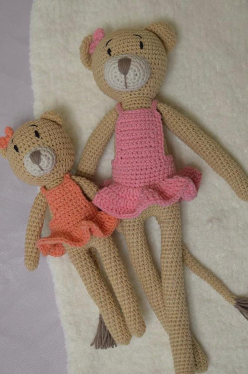May include: Two handmade crochet teddy bears. One bear wears a pink dress and the other wears an orange dress. Both bears have bows on their heads. The bears are tan with white and brown accents.