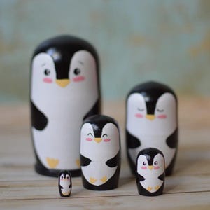 May include: Set of five black and white wooden nesting dolls shaped like penguins. The penguins have painted eyes, beaks, and feet. The largest penguin is about 10 cm tall.