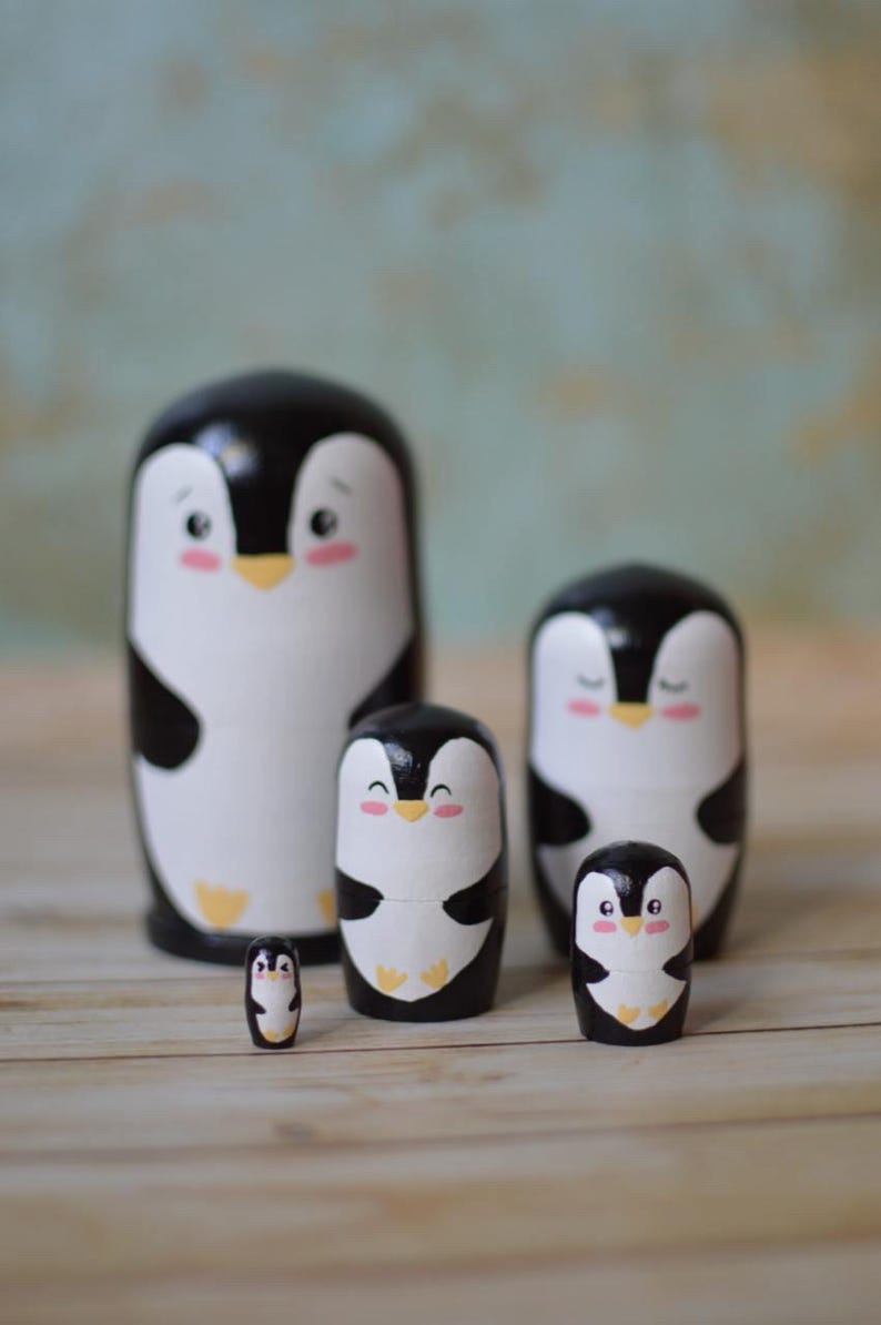 May include: Set of five black and white wooden nesting dolls shaped like penguins. The penguins have painted eyes, beaks, and feet. The largest penguin is about 10 cm tall.