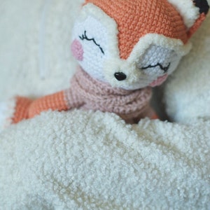 May include: A crocheted fox plush toy with closed eyes, a pink nose, and a pink scarf. The fox is being held by a person wearing a white fuzzy sweater.