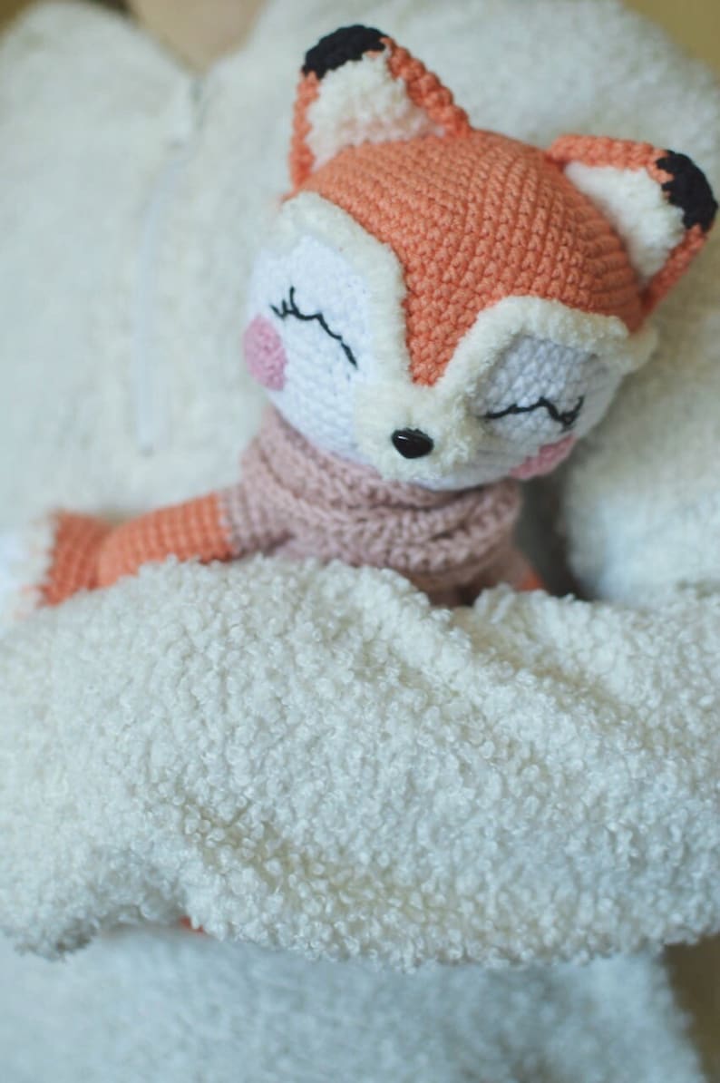 May include: A crocheted fox plush toy with closed eyes, a pink nose, and a pink scarf. The fox is being held by a person wearing a white fuzzy sweater.