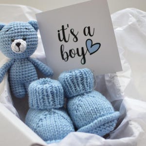Pode incluir: Um conjunto de presente para bebé numa caixa branca, com um ursinho de peluche azul de croché, dois botins de bebé azuis de malha e um cartão que diz "it's a boy" com um coração azul. Os artigos estão em papel de seda branco.