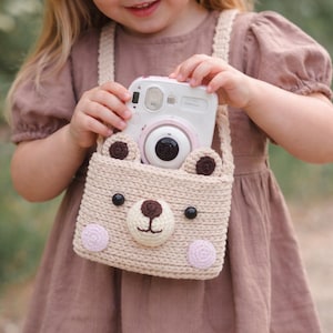 May include: A white and pink instant camera is held in a beige crocheted bear-shaped bag. The bag has brown ears, black eyes, and a brown nose. The bag has pink circles on each side of the bear's face. The camera has a black lens.