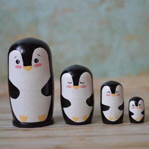 May include: A set of five black and white wooden nesting dolls shaped like penguins. The penguins have black eyes, pink cheeks, and yellow beaks. The largest penguin is about 4 inches tall.