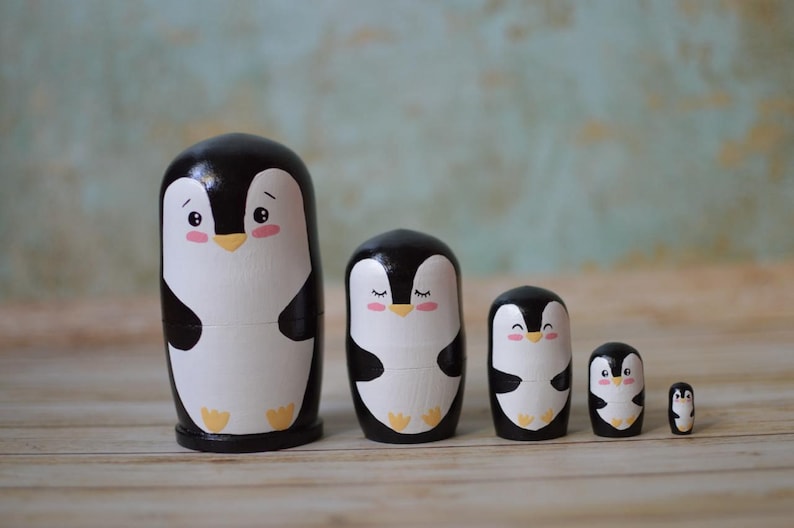 May include: A set of five black and white wooden nesting dolls shaped like penguins. The penguins have black eyes, pink cheeks, and yellow beaks. The largest penguin is about 4 inches tall.
