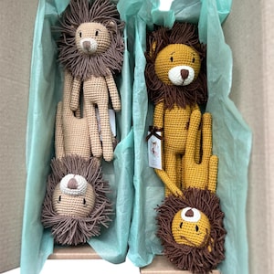 May include: A cardboard box filled with four crocheted lion plush toys. The lions have brown manes, tan bodies, and white and black facial features. The box is lined with light blue tissue paper, creating a soft, inviting presentation.