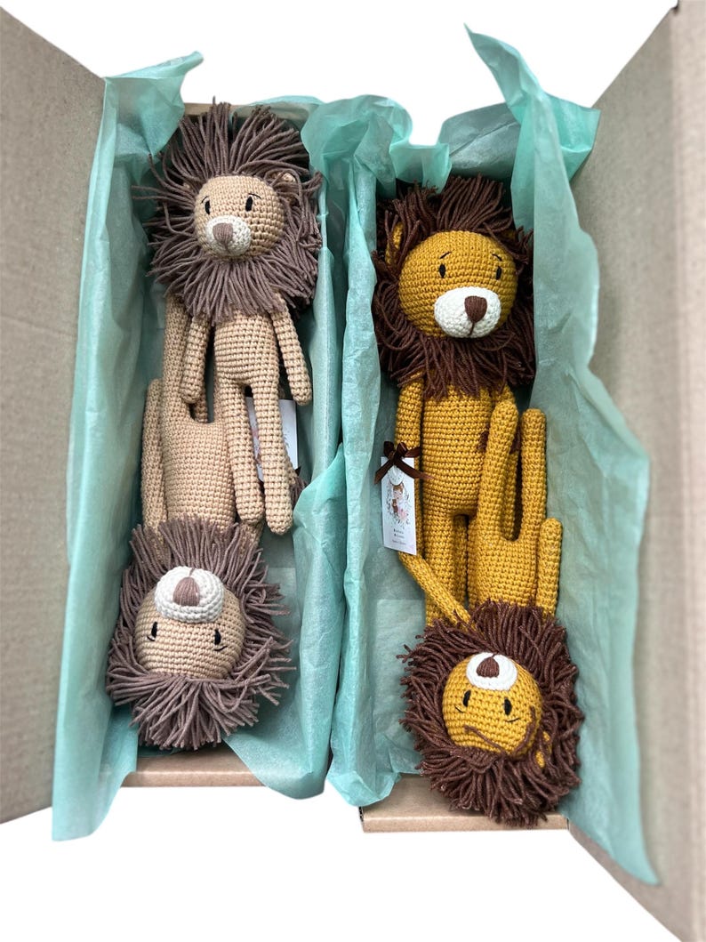 May include: A cardboard box filled with four crocheted lion plush toys. The lions have brown manes, tan bodies, and white and black facial features. The box is lined with light blue tissue paper, creating a soft, inviting presentation.