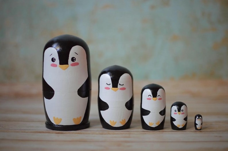 May include: A set of five black and white nesting dolls shaped like penguins. The penguins have black beaks, white bellies, and pink cheeks. The dolls are arranged in order of size, with the largest doll on the left and the smallest doll on the right.