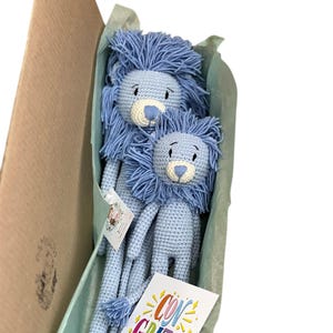 May include: Two blue crocheted lion stuffed animals in a brown cardboard box with a light blue tissue paper lining. The box also contains a card that says "Congrats!" in colorful letters.