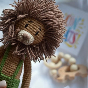 May include: A handmade crochet lion doll with a brown mane and a beige snout. The lion wears green overalls. A wooden teething toy and a card with colorful text are in the background. The doll is soft and cuddly.