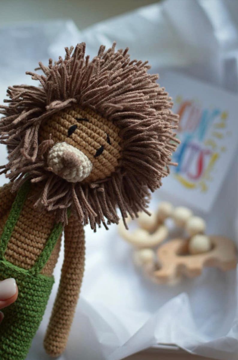 May include: A handmade crochet lion doll with a brown mane and a beige snout. The lion wears green overalls. A wooden teething toy and a card with colorful text are in the background. The doll is soft and cuddly.