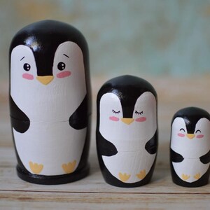 May include: A set of three nesting wooden penguin figurines. The largest penguin is black and white with a yellow beak and pink cheeks. The middle penguin is black and white with a yellow beak and closed eyes. The smallest penguin is black and white with a yellow beak and a smiling face.