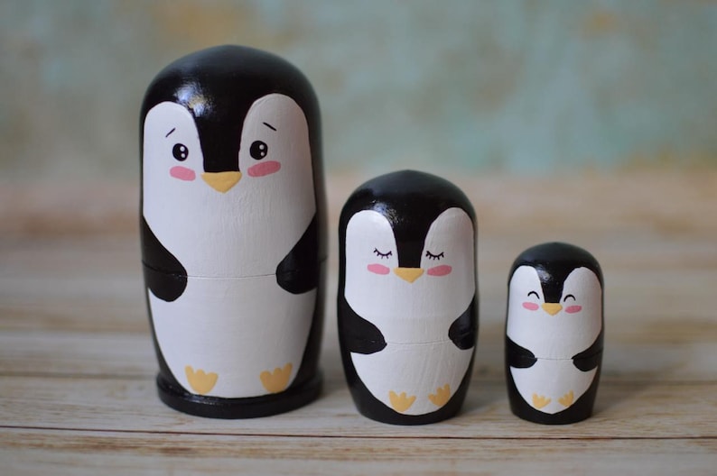 May include: A set of three nesting wooden penguin figurines. The largest penguin is black and white with a yellow beak and pink cheeks. The middle penguin is black and white with a yellow beak and closed eyes. The smallest penguin is black and white with a yellow beak and a smiling face.