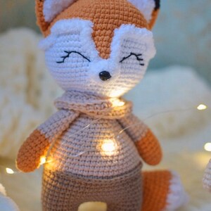 May include: A crocheted fox plush toy wearing a brown sweater and pants. The fox has a white face with black eyes and a black nose. The toy is sitting on a white fluffy surface with string lights.
