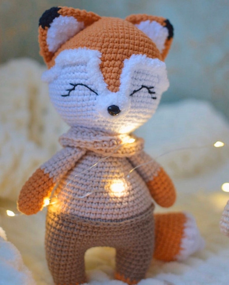 May include: A crocheted fox plush toy wearing a brown sweater and pants. The fox has a white face with black eyes and a black nose. The toy is sitting on a white fluffy surface with string lights.