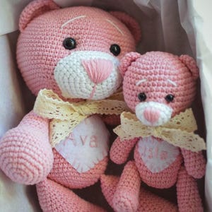 May include: Two pink crocheted teddy bears with white faces and black eyes. The larger bear has a white heart with the name "Ava" embroidered on it. The smaller bear has a white heart with the name "Isla" embroidered on it. Both bears are wearing a lace ribbon around their necks.