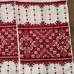 May include: A white fabric runner featuring intricate red cross-stitch embroidery. The design includes repeating geometric patterns, diamond shapes, and stylized floral motifs arranged in horizontal bands across the woven textile surface.