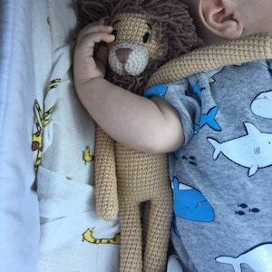May include: A crocheted lion stuffed animal with a brown mane and tan body. The lion is being held by a baby wearing a gray onesie with a blue shark pattern. The background includes a white and yellow giraffe print.