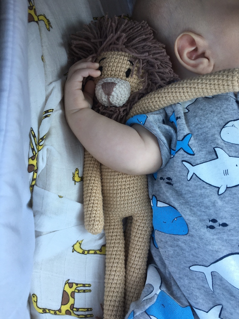 May include: A crocheted lion stuffed animal with a brown mane and tan body. The lion is being held by a baby wearing a gray onesie with a blue shark pattern. The background includes a white and yellow giraffe print.