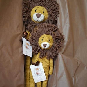 May include: Two handmade crochet lion stuffed animals in a brown box. The lions have yellow bodies, brown manes, and white and brown faces. Each lion has a tag with a floral illustration and a brown ribbon.