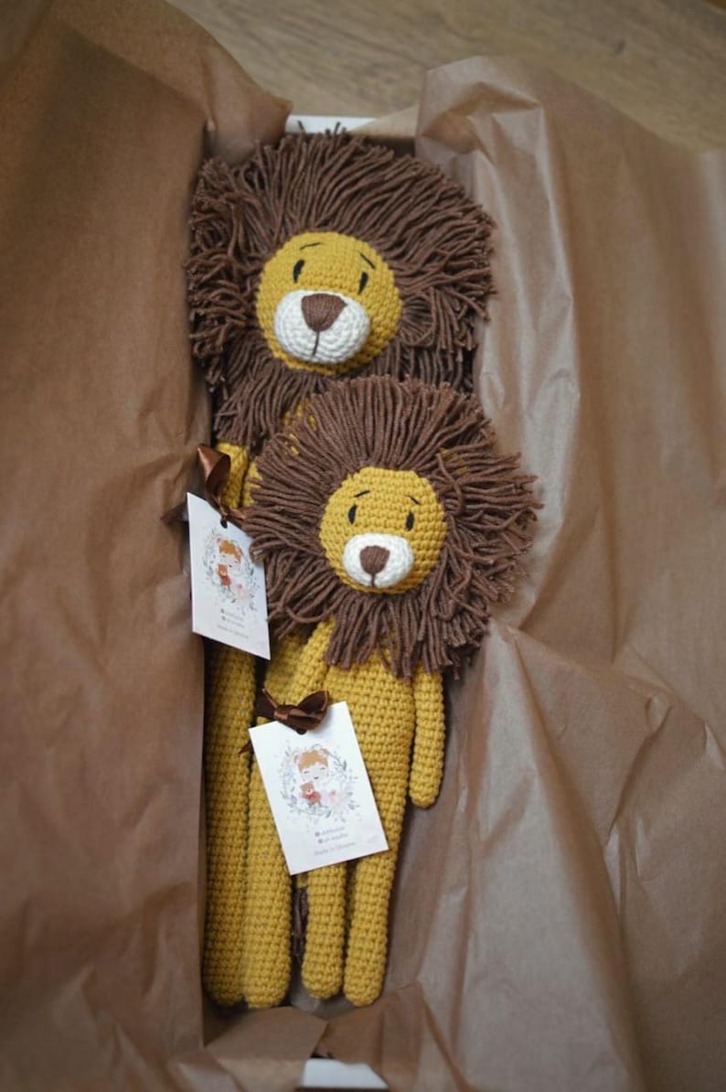 May include: Two handmade crochet lion stuffed animals in a brown box. The lions have yellow bodies, brown manes, and white and brown faces. Each lion has a tag with a floral illustration and a brown ribbon.