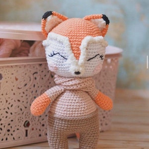 May include: A crocheted fox toy wearing a pink sweater and brown pants. The fox has orange fur, white facial features, and black eyes.