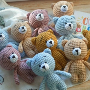Op de afbeelding: Een verzameling van tien gehaakte teddyberen in verschillende kleuren, waaronder blauw, roze, bruin en beige. De beren zitten allemaal op een wit stoffen oppervlak.