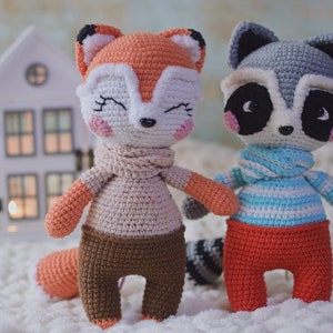 May include: Two crocheted stuffed animals, a fox and a raccoon, are sitting on a white surface. The fox is orange and white with a brown scarf and brown pants. The raccoon is gray with a blue and white striped scarf and red pants.