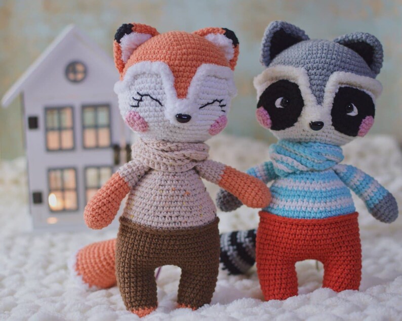 May include: Two crocheted stuffed animals, a fox and a raccoon, are sitting on a white surface. The fox is orange and white with a brown scarf and brown pants. The raccoon is gray with a blue and white striped scarf and red pants.