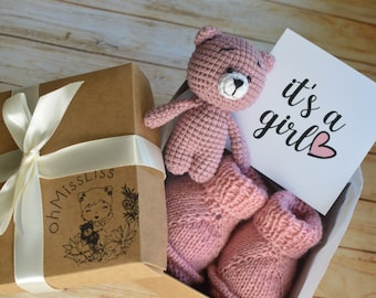 May include: A baby gift set in a brown box tied with a white ribbon. The box contains a pink crocheted teddy bear, a pair of pink knitted baby booties, and a card that reads "it's a girl" with a pink heart.