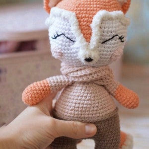 May include: A crocheted fox plush toy with closed eyes, wearing a brown sweater and pants. The fox is orange, white, and brown.