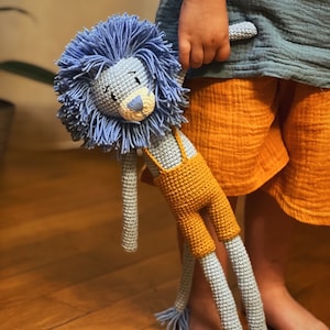 May include: A handmade crochet lion doll with a blue mane and gray body. The lion wears yellow overalls with suspenders. The doll is being held by a child wearing a blue shirt and orange shorts.