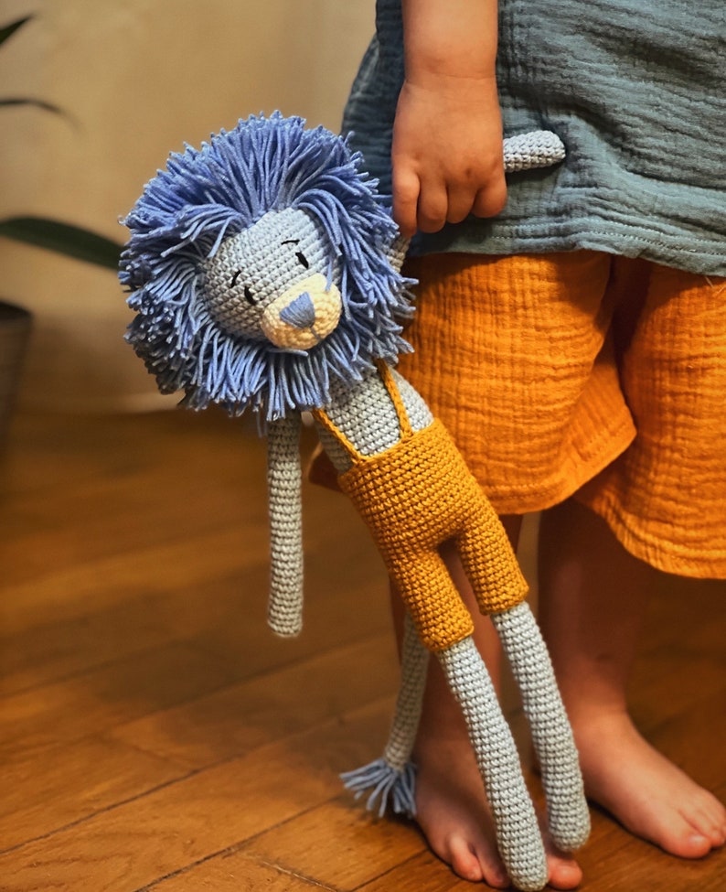 May include: A handmade crochet lion doll with a blue mane and gray body. The lion wears yellow overalls with suspenders. The doll is being held by a child wearing a blue shirt and orange shorts.