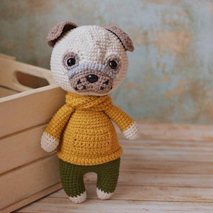Puede incluir: Un pug de crochet con un suéter amarillo y pantalones verdes. El pug está de pie sobre una superficie de madera.