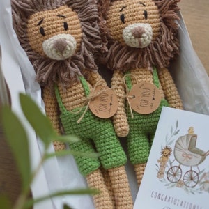 May include: Two handmade crochet lion dolls with brown manes and tan bodies, wearing green overalls. Each doll has a tag that says "FOR YOU". A card with a baby carriage illustration and the word "CONGRATULATIONS!" is also included.