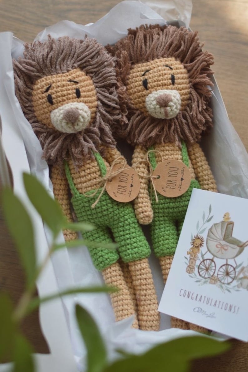 May include: Two handmade crochet lion dolls with brown manes and tan bodies, wearing green overalls. Each doll has a tag that says "FOR YOU". A card with a baby carriage illustration and the word "CONGRATULATIONS!" is also included.