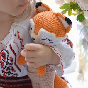 May include: A young girl holds a crocheted fox plush toy. The fox is orange and white with black eyes and a pink nose. The girl is wearing a white shirt with red and black embroidery.
