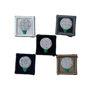May include: Five square embroidered patches featuring a golf ball design. Each patch has a white golf ball on a green tee, set against a different colored background: white, black, navy, gray, and tan. The patches are bordered with matching thread.