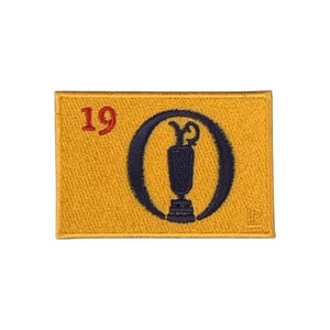 Puede incluir: Un parche rectangular bordado con fondo amarillo dorado. Presenta el número 19 en rojo en la esquina superior izquierda y un emblema circular azul oscuro en el centro que contiene un icono de trofeo estilizado. Un pequeño logotipo está en la parte inferior derecha.