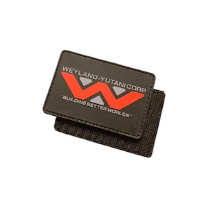 Puede incluir: Un parche rectangular negro con el texto "WEYLAND-YUTANI CORP" y "BUILDING BETTER WORLDS" en blanco. El logotipo presenta una forma de "W" roja con acentos grises. El parche está adherido a una tela gris oscuro texturizada.
