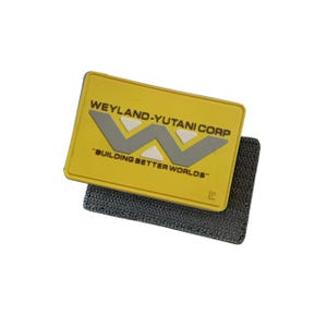 May include: A yellow rectangular patch with the text "WEYLAND-YUTANI CORP" and "BUILDING BETTER WORLDS" in white and gray. The patch has a gray and white logo and is attached to a black, textured fabric.