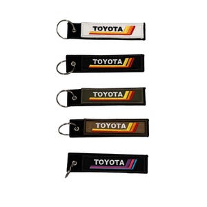 May include: Five embroidered keychains with the word "TOYOTA" in white text. Each keychain has a different colored background: white, black, olive green, brown, and black. Each keychain has a metal ring for attaching keys.