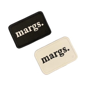 May include: Two rectangular patches with rounded corners. One patch is black with white text that reads "margs." The other patch is off-white with black text that reads "margs." Both patches have a small logo in the corner.