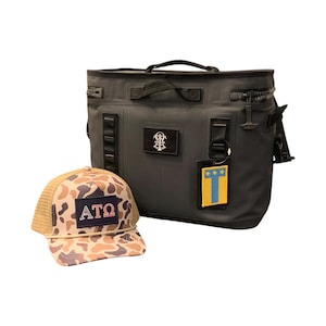 Op de afbeelding: Een donkergrijze geïsoleerde koeltas met een zwart handvat en rits, naast een bruin en camouflage truckerpet. De pet heeft een patch met de letters "ATO". De koeltas heeft een patch met een wapen en een geel en blauw label.