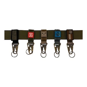 Könnte beinhalten: Ein dunkelgrüner Gürtel mit fünf Schlüsselanhängern. Jeder Schlüsselanhänger hat ein anderes Patch: eine amerikanische Flagge, ein rotes Patch mit "LP PATCH", ein Patch mit gekreuzten Lacrosse-Schlägern, ein blaues Friedenszeichen und ein Patch mit einem kreisförmigen Design.