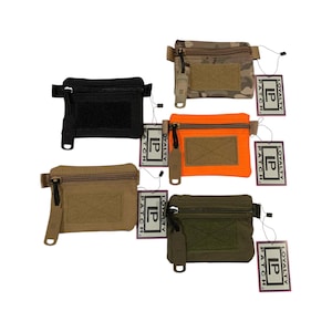 Könnte beinhalten: Eine Sammlung kleiner, rechteckiger Taschen in verschiedenen Farben: Schwarz, Beige, Orange, Tarnmuster und Olivgrün. Jede Tasche hat einen Reißverschluss, einen Klettverschluss und ein kleines Etikett mit der Aufschrift "LOYALTY PATCH".